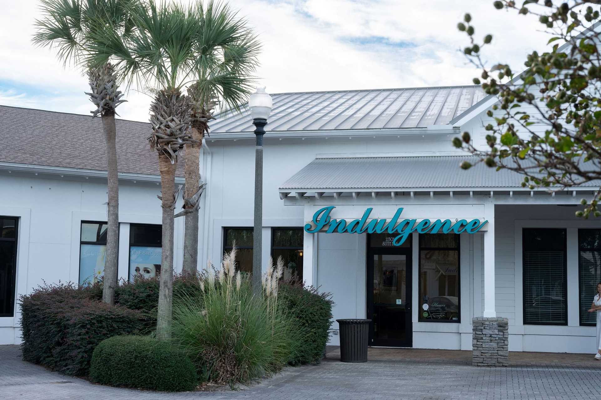 Storefront with "Indulgence" sign, palm trees, and bushes in front of a white building.